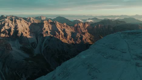 Vista-Aérea-Escénica-De-La-Escarpada-Cordillera-Dolomita-En-Italia-Al-Amanecer,-Con-Un-Valle-Cubierto-De-Nieve-Y-Laderas-En-Primer-Plano