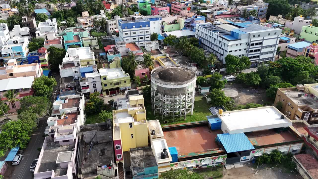 An expansive, high-angle aerial shot captures a prominent, large circular water tank at the epicenter of a dense Indian residential community. The composition highlights the intricate urban sprawl