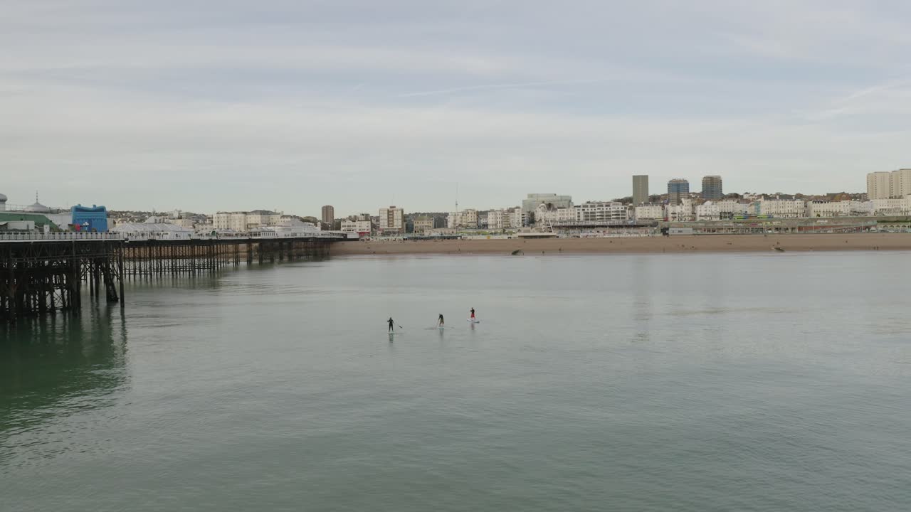 tres personas en tablas de paddle rumbo a la playa en brightonm, gaviotas volando y drones panoramizando a la derecha