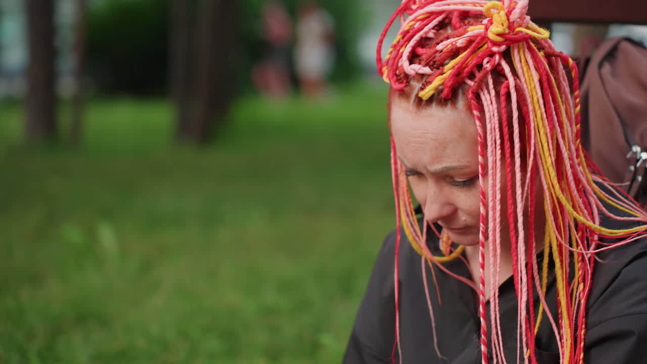 Primer plano de una mujer blanca con rastas de colores llorando, con la mano cubriendo la boca y luego la cara, bokeh suave de árboles detrás, captura visceral de la tristeza, expresión cruda, rastros de lágrimas y aliento tembloroso