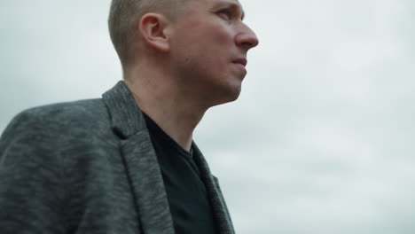 a close view of an aged white man wearing a grey suit and a black polo shirt, turning back with a focused expression, with an overcast sky in view