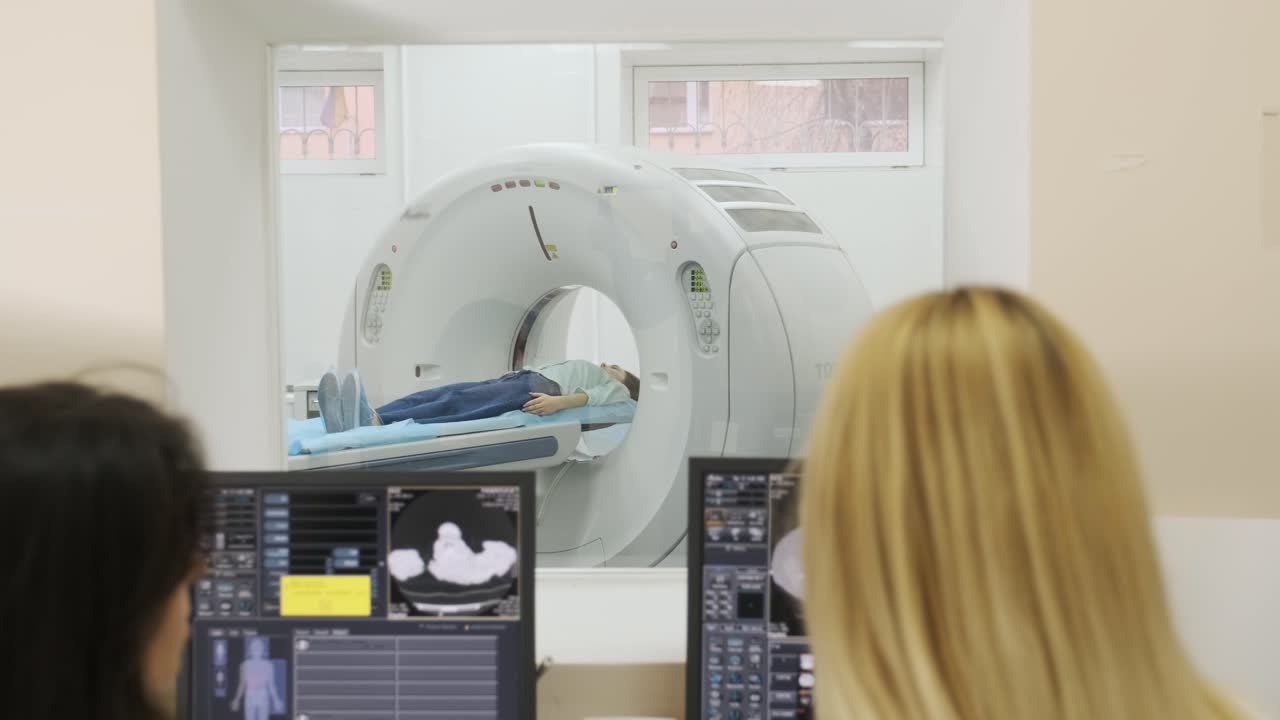 dos radiólogos calificados examinan a una paciente femenina que está acostada bajo un escáner de ct o mri. pantalla de computadora con imagen de diagnóstico de resultado. vista a través de una ventana de vidrio en una clínica moderna.