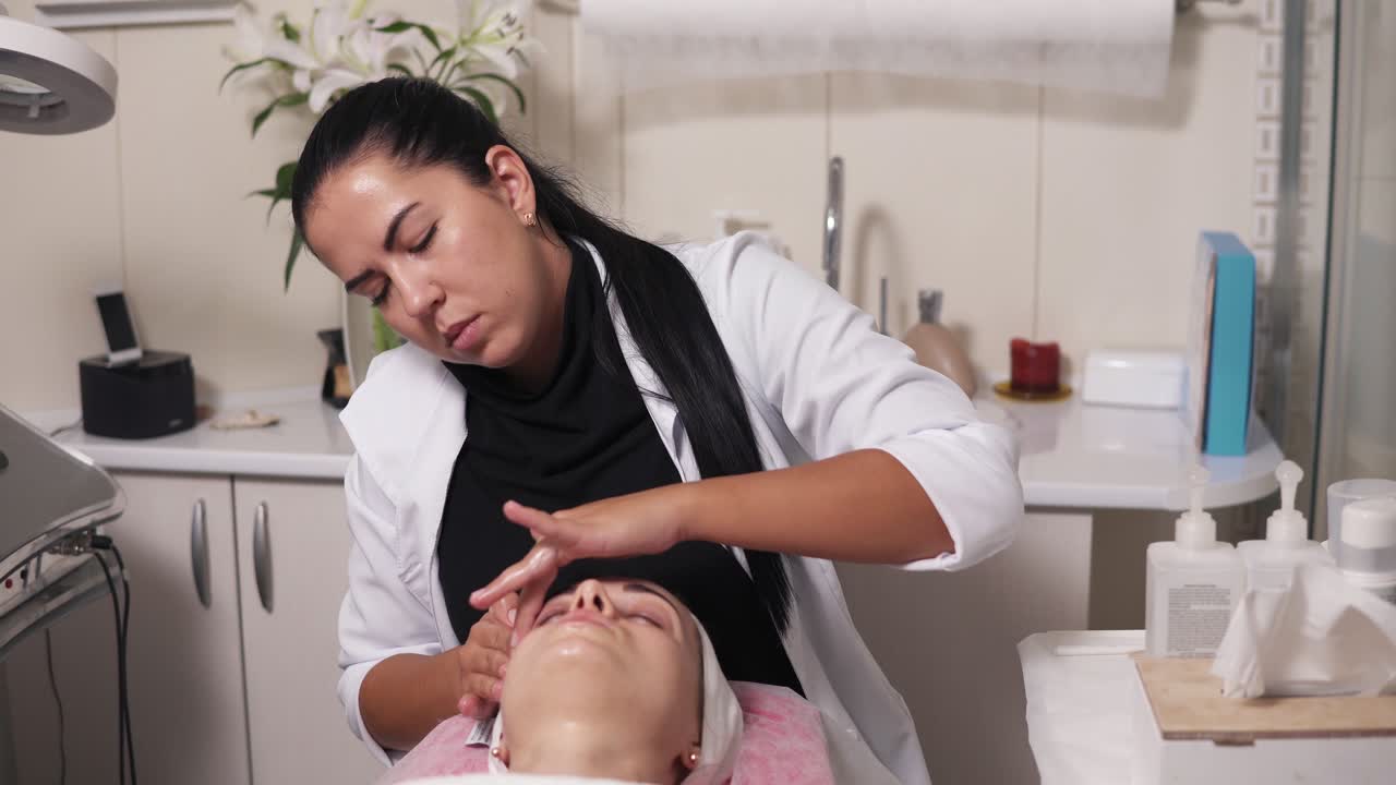 cosmetólogo profesional está haciendo masaje facial en el salón de spa. mujer joven está hablando con su cosmetólogo mientras está acostada en el sofá durante el procedimiento facial cosmético. 4k tiro