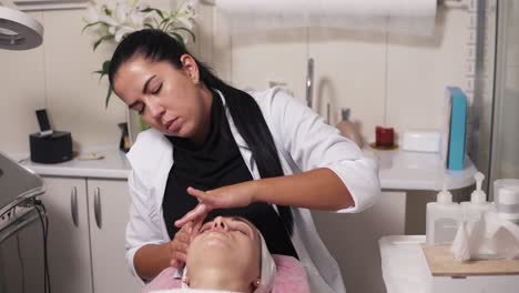 professional cosmetologist is making face massage in spa salon. young woman is talking to her cosmetologist while lying on the couch during cosmetic face procedure. 4k shot