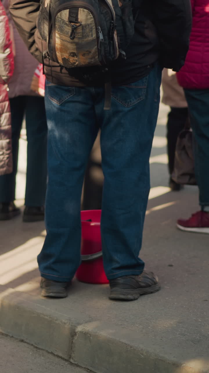 adulto con zaino in piedi sul marciapiede, jeans in denim e giacca scura, secchio rosso per le donazioni sul selciato, folla di pedoni in attesa, luce del sole mattutina che proietta lunghe ombre, volontario e pendolare
