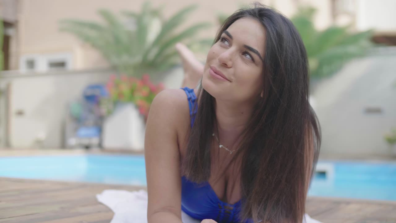 hermosa mujer morena de pelo largo tomando el sol y sonriendo junto a la piscina en el balneario