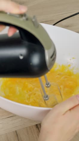 hand-held mixer whisks together bright yellow egg yolks with powdered sugar in a bowl, vertical kitchen close-up shot during dessert preparation, camera static, making tiramisu