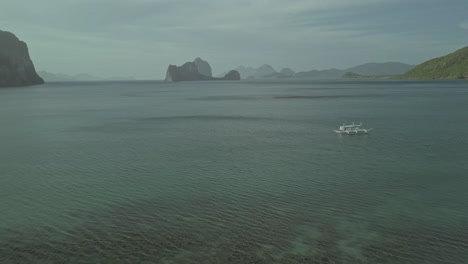 Catamaran-boat-in-tropical-water-by-cliffy-islands-in-Philippines,-aerial-pull-out