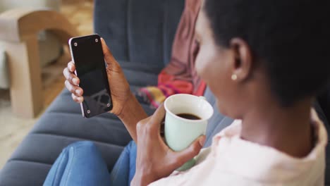 Feliz-Mujer-Afroamericana-Sentada-En-Un-Sofá-En-La-Sala-De-Estar,-Usando-Un-Teléfono-Inteligente-Con-Espacio-Para-Copiar