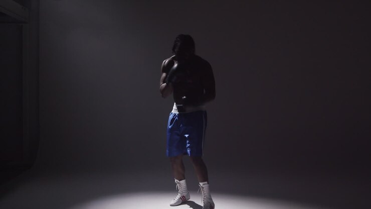 Silhouetted Studio Shot With Low Key Lighting Of Boxer Putting Up Gloves Ready To Fight Standing In Pool Of Light Against Grey Background