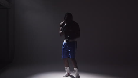 Silhouetted-Studio-Shot-With-Low-Key-Lighting-Of-Boxer-Putting-Up-Gloves-Ready-To-Fight-Standing-In-Pool-Of-Light-Against-Grey-Background