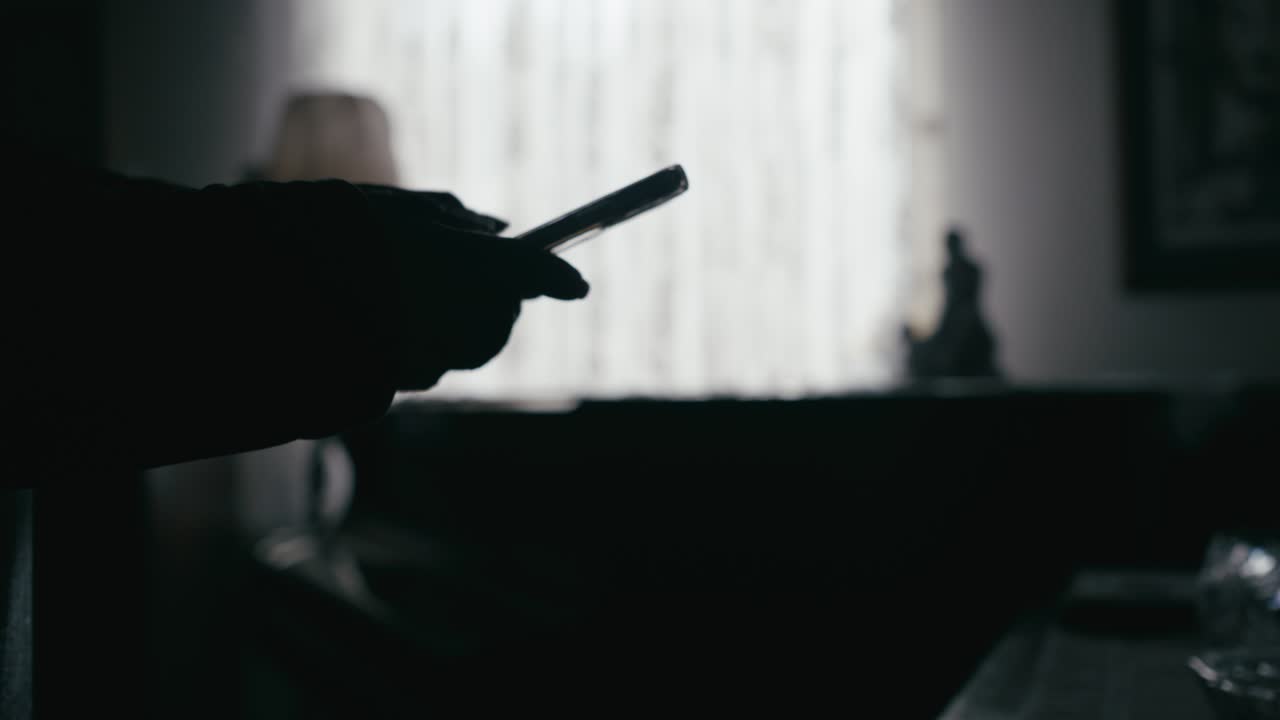 Silhouetted man texts message in Dark room backlit by veiled curtains