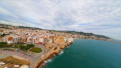 FPV-aerial-drone-flying-along-the-coast-of-Sitges,-Spain-with-a-view-of-Sant-Bartomeu-i-Santa-Tecla-church