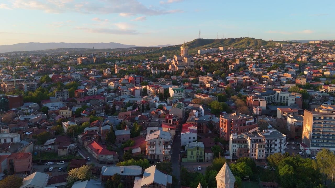 Luftpanorama af det historiske Avlabari-distrikt i Tbilisi, Georgien, der viser traditionelle røde tegltage og den majestætiske Hellig Treenigheds Katedral (Sameba), der står på bakken ved solnedgang.