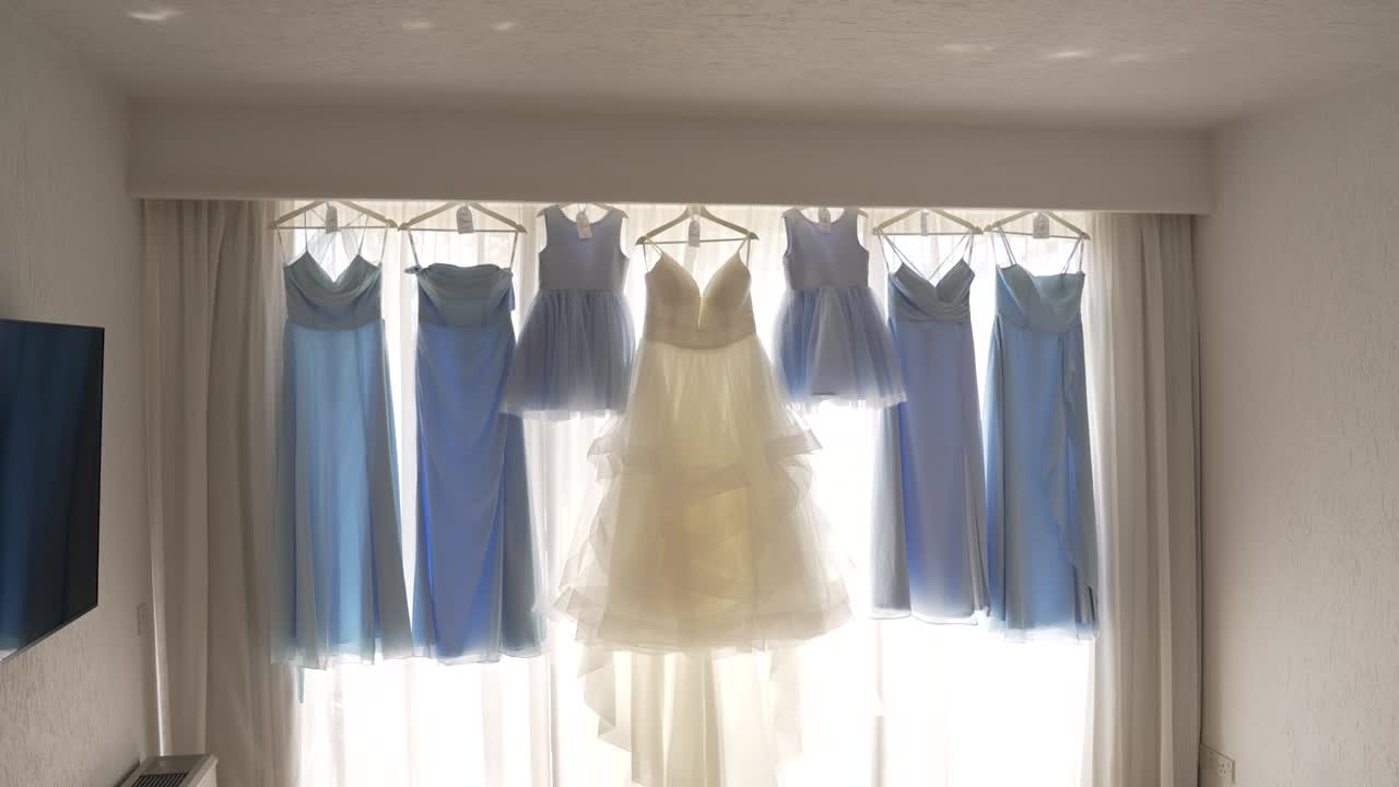 A stunning display of blue and white dresses is hung in front of a bright window, highlighting their delicate fabric and elegant designs. Perfect for a special occasion