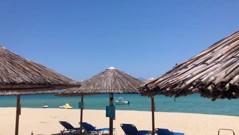 Vista-Panorámica-De-La-Playa-De-Sarti-En-Sithonia,-Halkidiki-En-Cámara-Lenta-Con-Vistas-A-Las-Tumbonas-Azules-Vacías-Bajo-Sombrillas-Tiki-Bar-Y-Vista-Frente-A-La-Playa-En-El-Mar-Mediterráneo-Cian-Con-Motos-Acuáticas