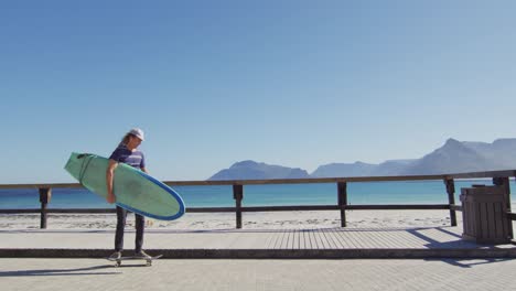 Video-Eines-Kaukasischen-Mannes-Mit-Dreadlocks,-Der-Auf-Der-Sonnigen-Strandpromenade-Skateboard-Fährt-Und-Ein-Surfbrett-Trägt