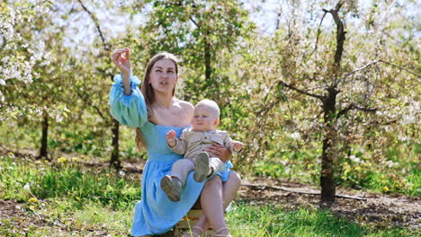 long-haired woman in blue dress sits outdoor holding her baby. mother shows a flower to her son in the garden.
