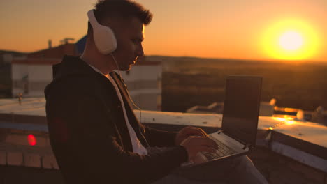 Ein-Freiberuflicher-Mann-Mit-Kopfhörern-Steht-Bei-Sonnenuntergang-Auf-Dem-Dach-Und-Schreibt-Codeseiten-Auf-Der-Tastatur.-Kleines-Geschäft.-Höre-Musik-Und-Arbeite-Am-Computer-Und-Genieße-Die-Schöne-Aussicht-Vom-Dach.