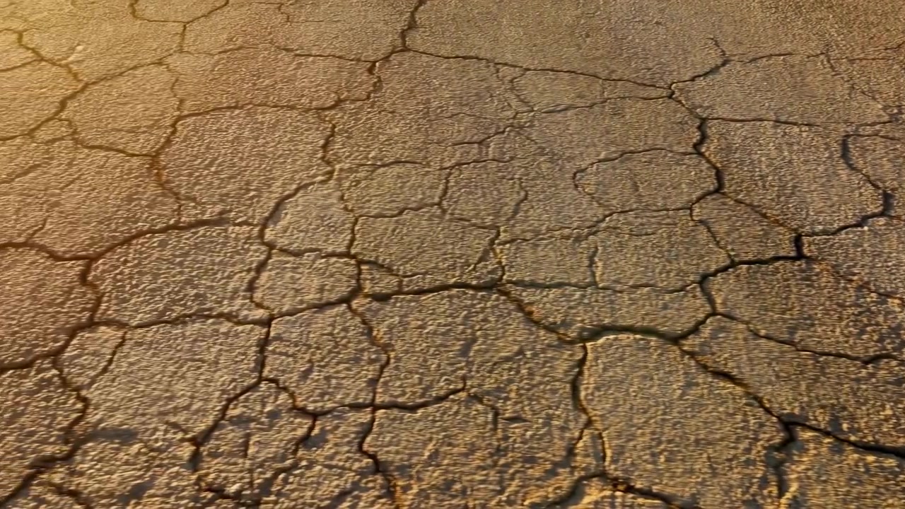 Primo piano dal basso di terra secca e screpolata illuminata dal sole che riempie l'inquadratura, mostrando placche poligonali irregolari e fessure poco profonde con illuminazione laterale calda e una superficie texturizzata.