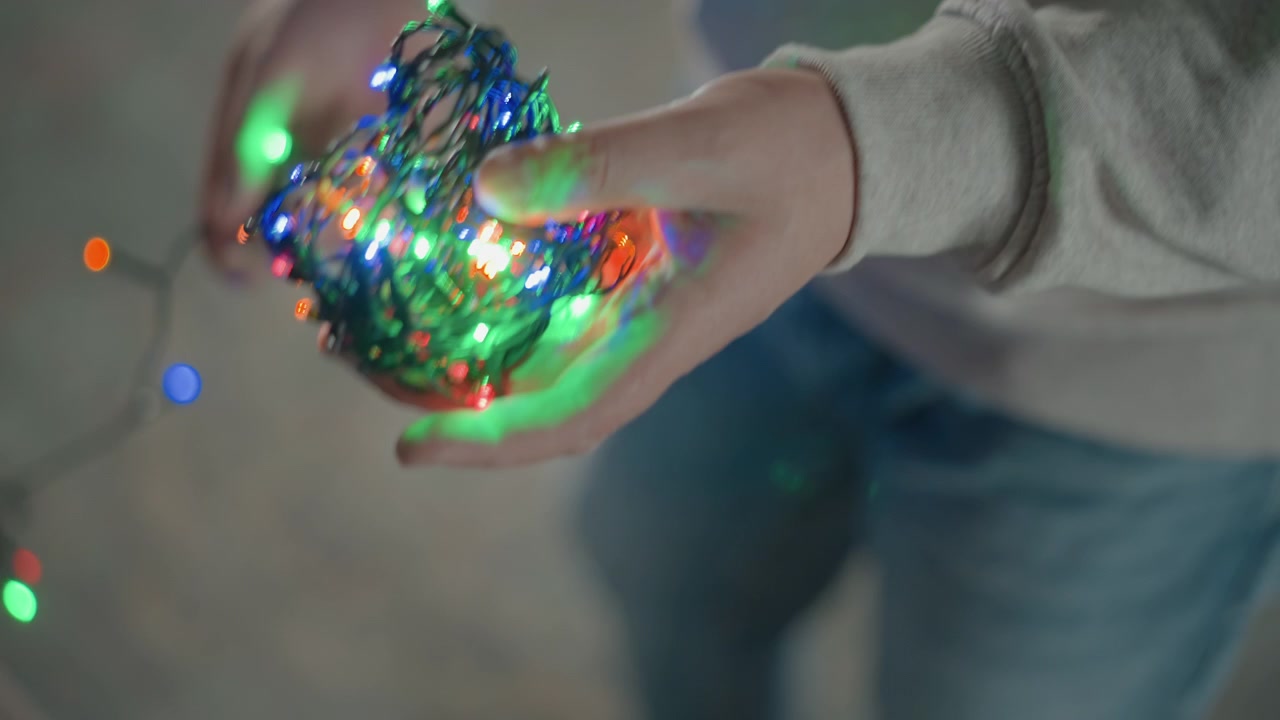 Close slightly elevated view of hands untangling and spreading compact bundle of glowing multicolored LED string lights, with visible finger movement, soft indoor light, cozy mood and festive decor