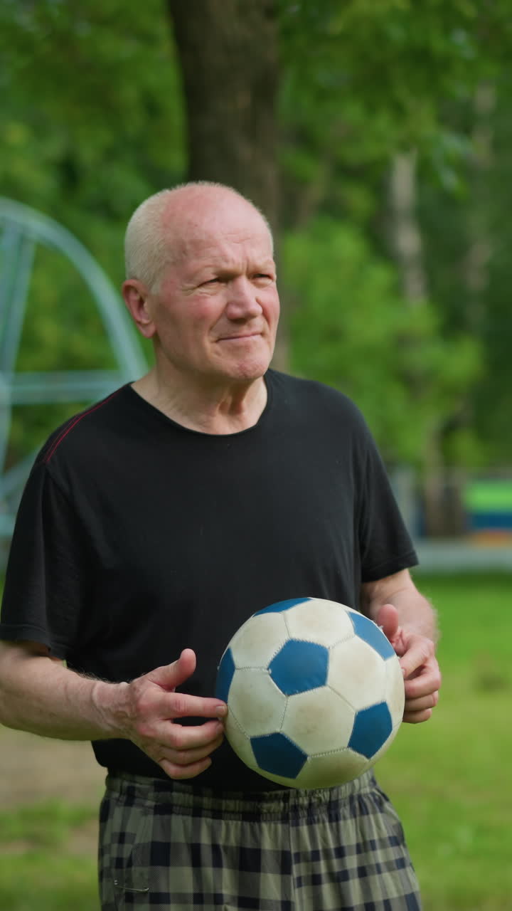 An elderly man, focused and contemplative, rolls a soccer ball in his hand while looking down, with a background view of goal post and outdoor equipment in the background