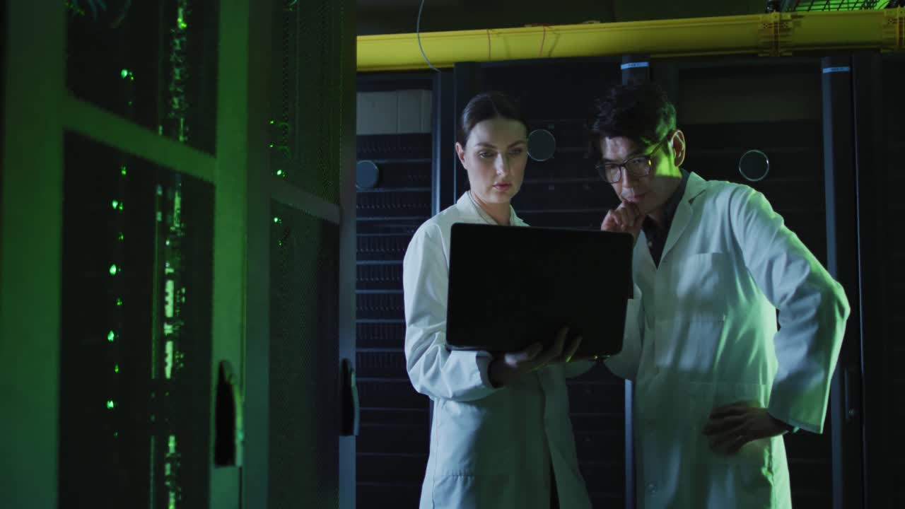 Diverse female and male it technicians in lab coats using laptop checking computer server