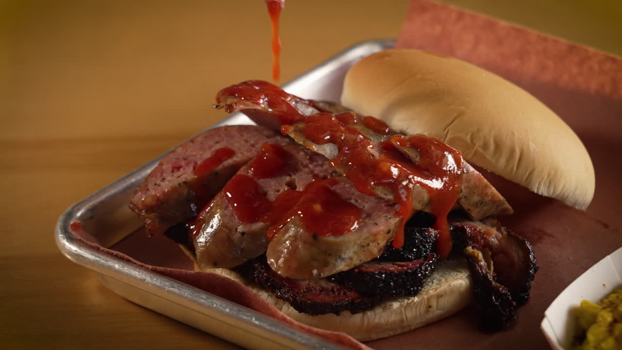 Salsa barbecue rossa versata su panino con salsiccia affumicata tradizionale del Texas e brisket a fette, ripresa ravvicinata in slow motion 4K.