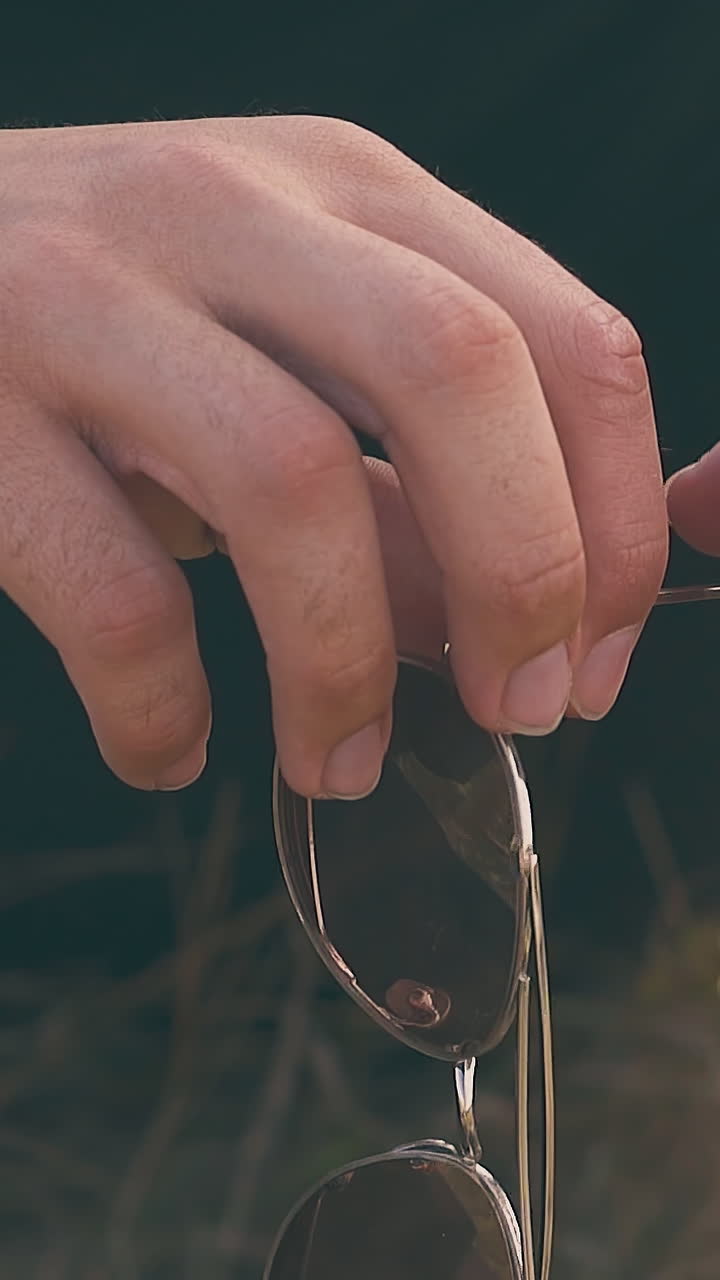 man hands hold fashionable sunglasses sitting on brown meadow lit by autumn sun slow motion extreme close view