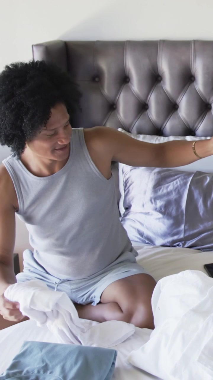 Video verticale: Un uomo con canotta grigia rifà il letto in camera da letto, raggiungendo il cesto della biancheria e spostando il cellulare.