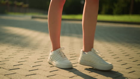 leg view of white lady wearing white canvas shoes walking along interlocked path, shadow cast on ground, with blur background featuring greenery and soft light glow