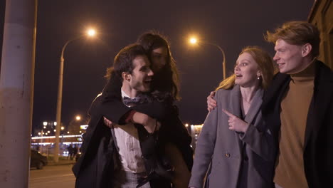 two couples of friends dressed in elegant clothes walking in the street street while talking and laughing after new year's party, one of the girls is perched on her boyfriend's back