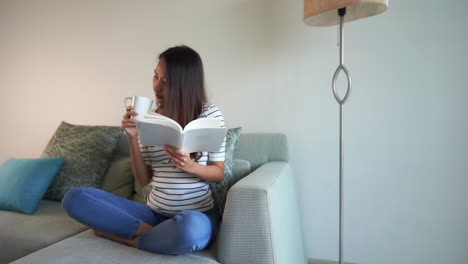 Sonriente-Mujer-Asiática-Leyendo-Un-Libro-Y-Bebiendo-Té-En-El-Sofá-En-La-Sala-De-Estar-De-La-Casa-Familiar