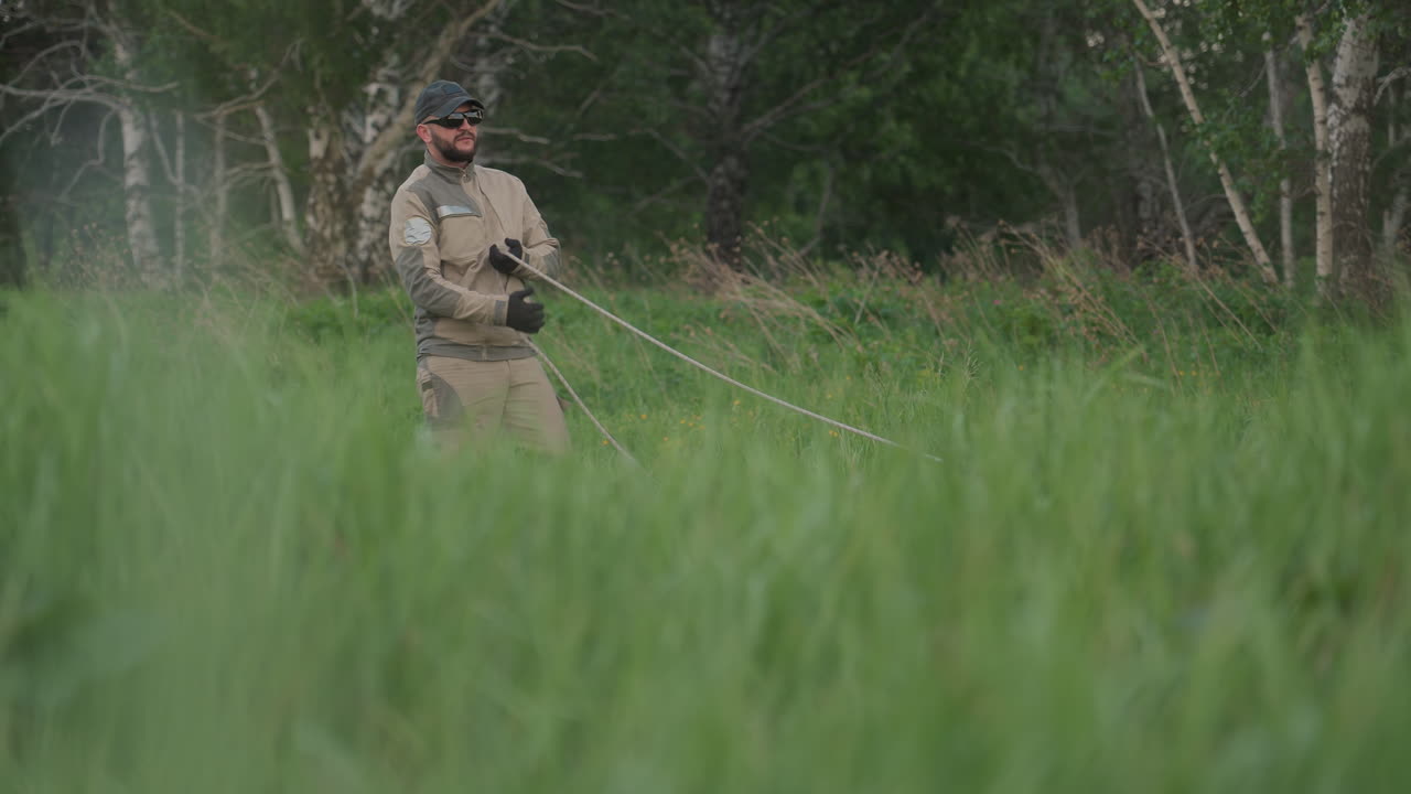 man in cap and gloves standing in tall grass pulling long rope during hot air balloon preflight setup in rural field at sunset, preparing lines for inflation