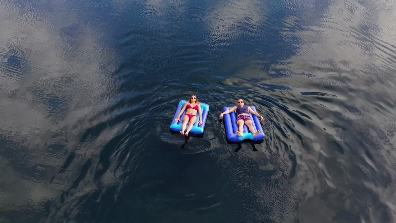 Two women enjoy summer, floating and sunbathing on inflatable mattresses in a calm river, surrounded by lush vegetation and reflections of the sky