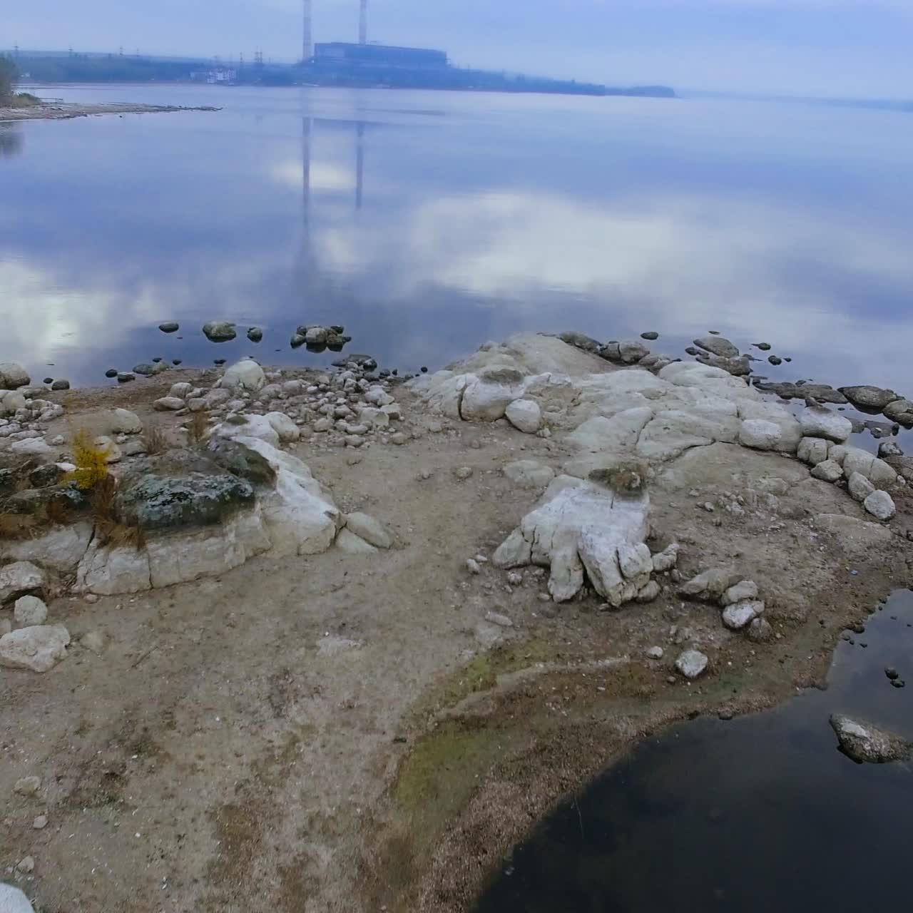 Rocky bank of the river with no plants growing on. Grey clouds reflecting in the smooth surface of the water. Pipes of industrial factory at backdrop