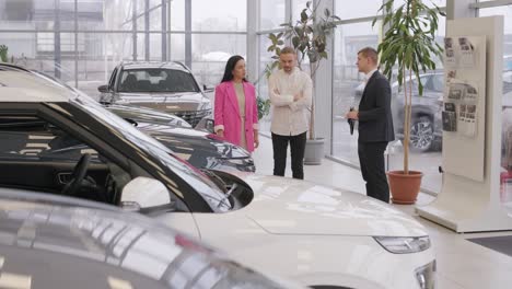 young car salesman showing to young couple new automobile at dealership salon.