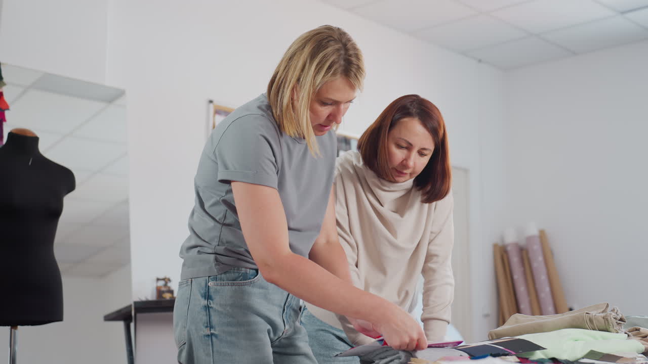 Seamstress mentoring apprentice in bright fashion studio explaining fabric designs while standing by table filled with colorful materials samples sketches rolls of fabric and inspiring workspace