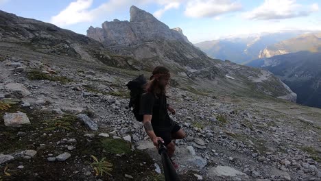 a young, fit man with long hair and a large backpack is jogging and running carefully down a path in themountains full of rocks and stones