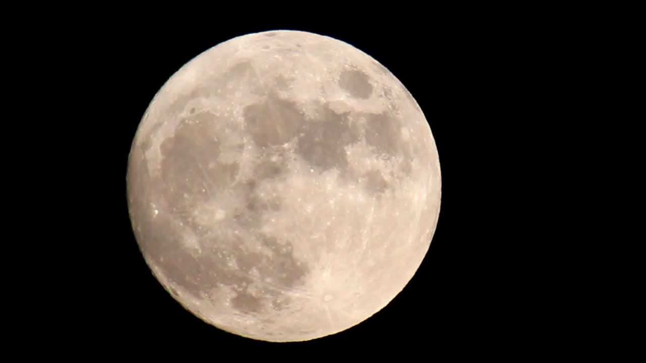 Com zoom em uma lua cheia brilhante amarelo-alaranjada que preenche o quadro, com o zoom diminuindo até a lua estar no centro do quadro, ligeiramente deslocada para a esquerda