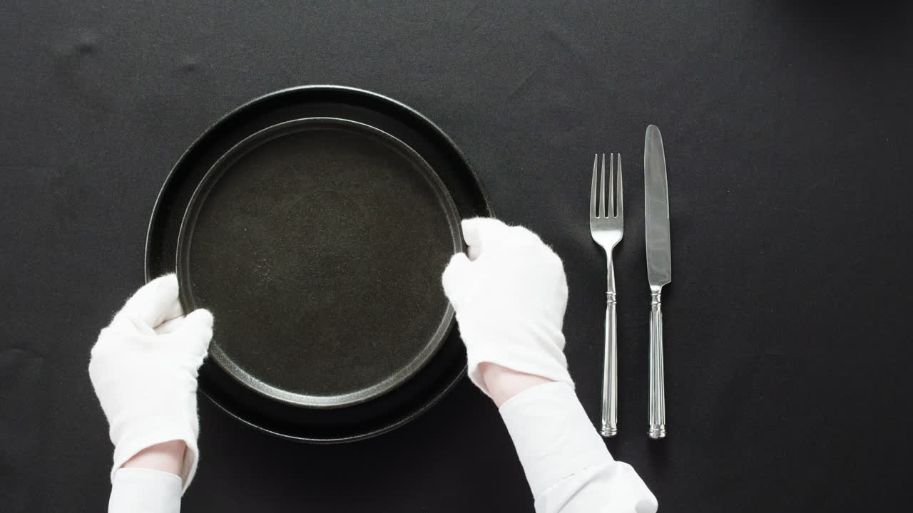 Professional table setting with white gloves. Plates, fork, knife, and glass arranged on a white table.