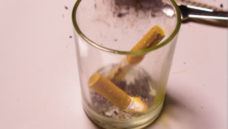 close up of a tiny cheap glass placed on a white table inside a home person smoking filtered cigarette by themselves lonely at home depressed about life flicking ash inside the cup with mess around