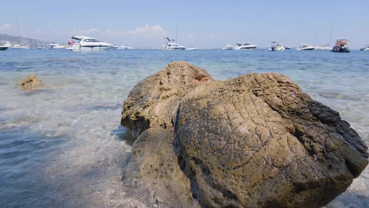 Nærbillede af store forvitrede klipper i lavt, klart vand med både ankret op i baggrunden, Ile Sainte-Marguerite, Frankrig