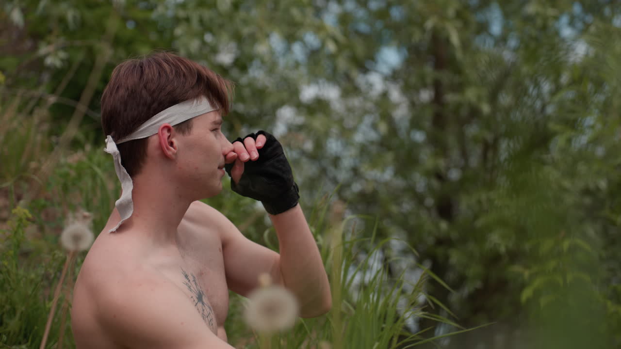 Side view of young man seated near riverbank under cloudy daylight, wearing headband and gloves, rubbing nose thoughtfully as lush green trees surround calm water in serene outdoor setting