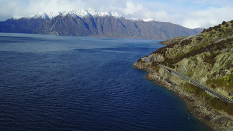 Aerial-Tracking-shot-of-RV-driving-on-a-coastal-road-that-then-pans-to-a-snowy-mountain-range