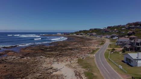 Sudáfrica-Aerea-Dron-Cinemático-Playa-Pequeño-Surf-Pueblo-Jardín-Ruta-Quiet-Bay-Jeffreys-Bay-Casas-Y-Edificios-Olas-Rompiendo-En-Arrecife-Gente-Caminando-Calle-A-última-Hora-De-La-Mañana-Tarde-Adelante-Movimiento