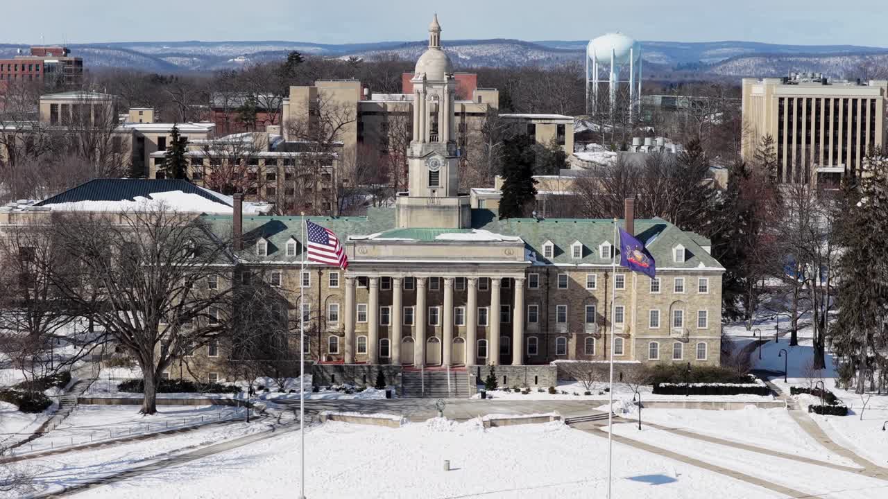 Dronebillede af Penn State Universitets Old Main-bygning dækket af frisk sne, med amerikanske flag, der vajer foran det historiske universitetslandemærke på en lys vinterdag. Vidvinkelbillede
