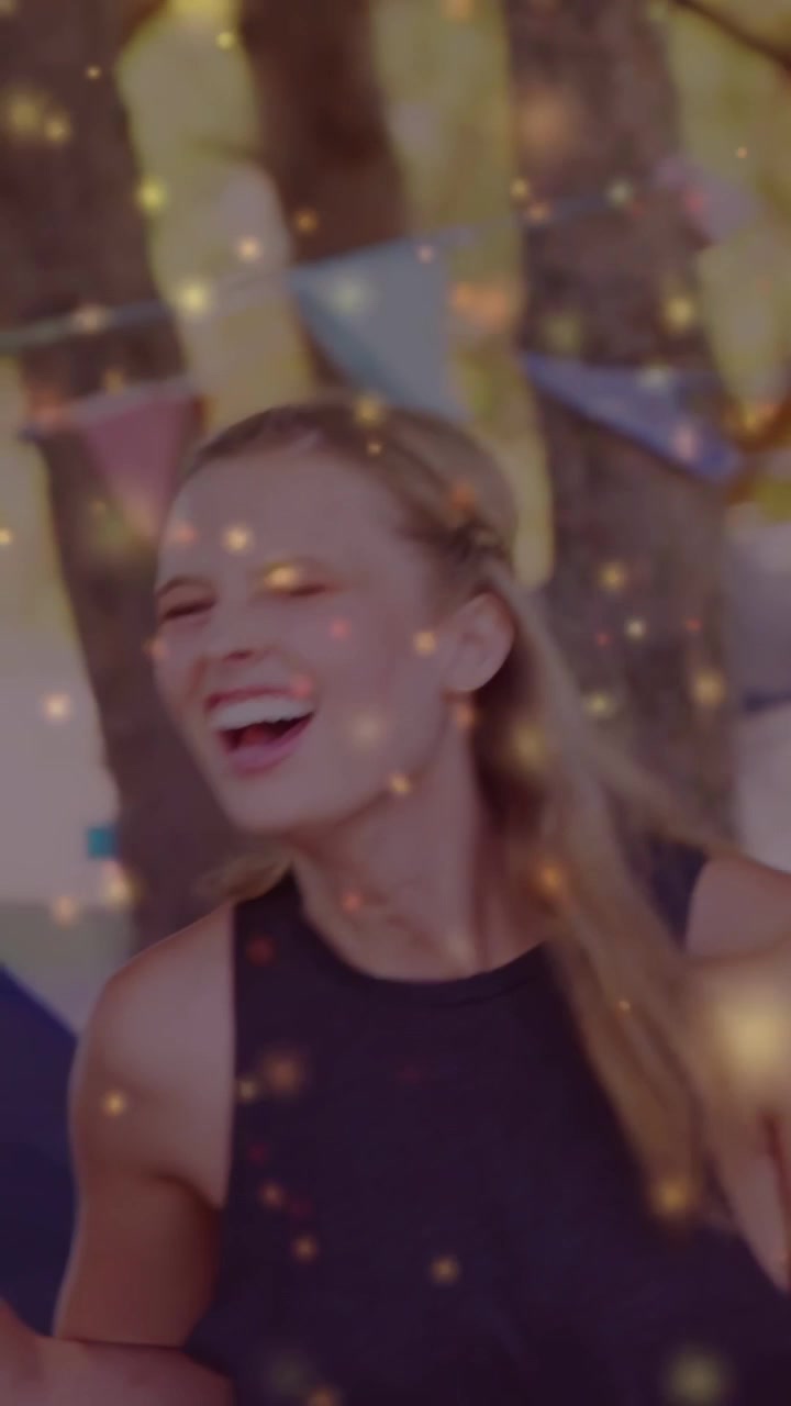 Video verticale: Donna in canotta che si sporge mentre un uomo si avvicina, ridendo a un evento, con bokeh dorato sul gruppo.
