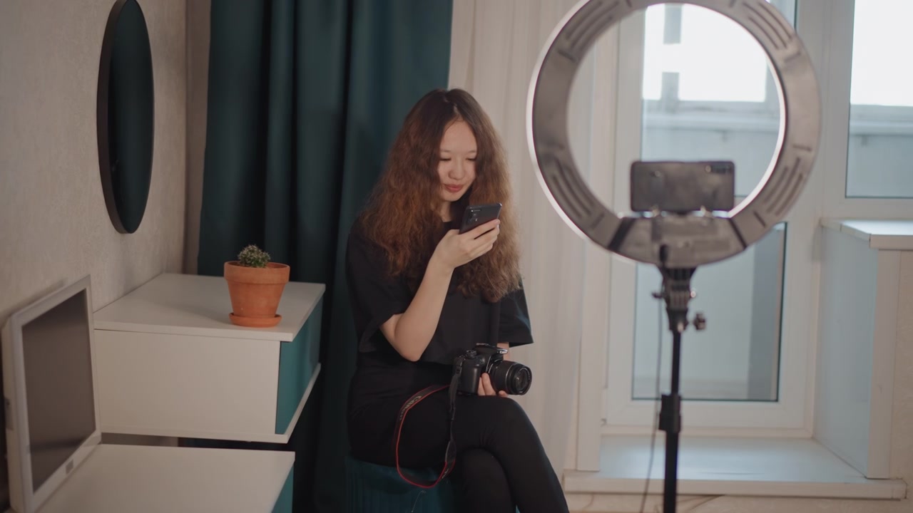 Giovane donna in abbigliamento casual scuro parla al suo smartphone vicino a una ring light e una fotocamera in un accogliente angolo di studio casalingo, con luce naturale dalla finestra, decorazioni con piante, espressione concentrata e posa creativa