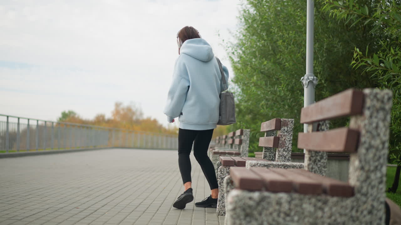 Pretty lady walking in park to sit on concrete bench, listening to music with headphones, peaceful outdoor environment with benches, green leaves, and serene atmosphere